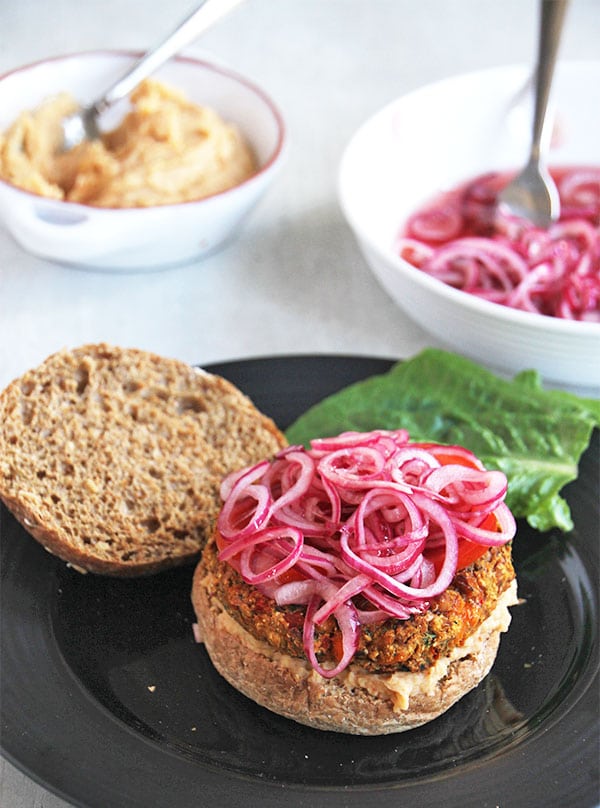 Mediterranean veggie burger on black plate with bowl of white bean spread and bowl of pickled red onions.