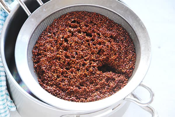rinsed red quinoa in mesh strainer over stainless steel pot.