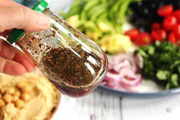 oil free vinaigrette being shaken in jar with hummus and Greek salad toppings in background.