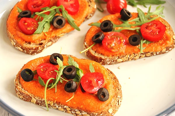 Red pepper Romesco sauce on bruschetta with black olives, tomatoes and arugula on white and blue plate.