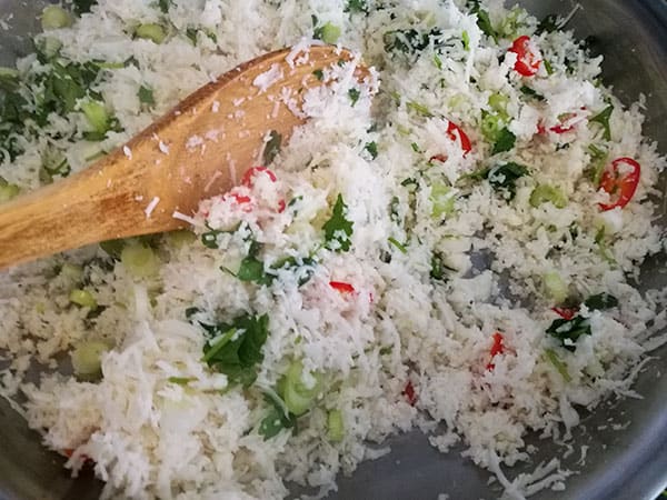 Stirring cauliflower rice with red chilis, scallions and fresh coriander with a wooden spoon in a silver skillet.