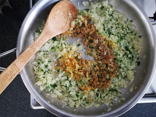 Turmeric, coriander, cumin and cayenne are added to diced onions and jalapenos in steel pan with wooden spoon.