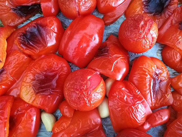 Red peppers, tomatoes and garlic are roasted in the oven on a baking tray lined with parchment paper.