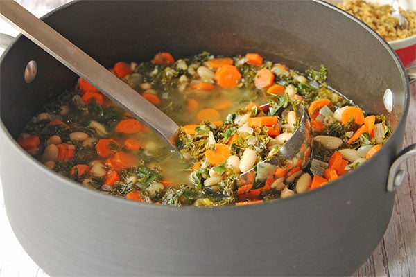 Italian white bean soup with carrots, kale and fresh herbs in black soup pot with ladle lifting soup.