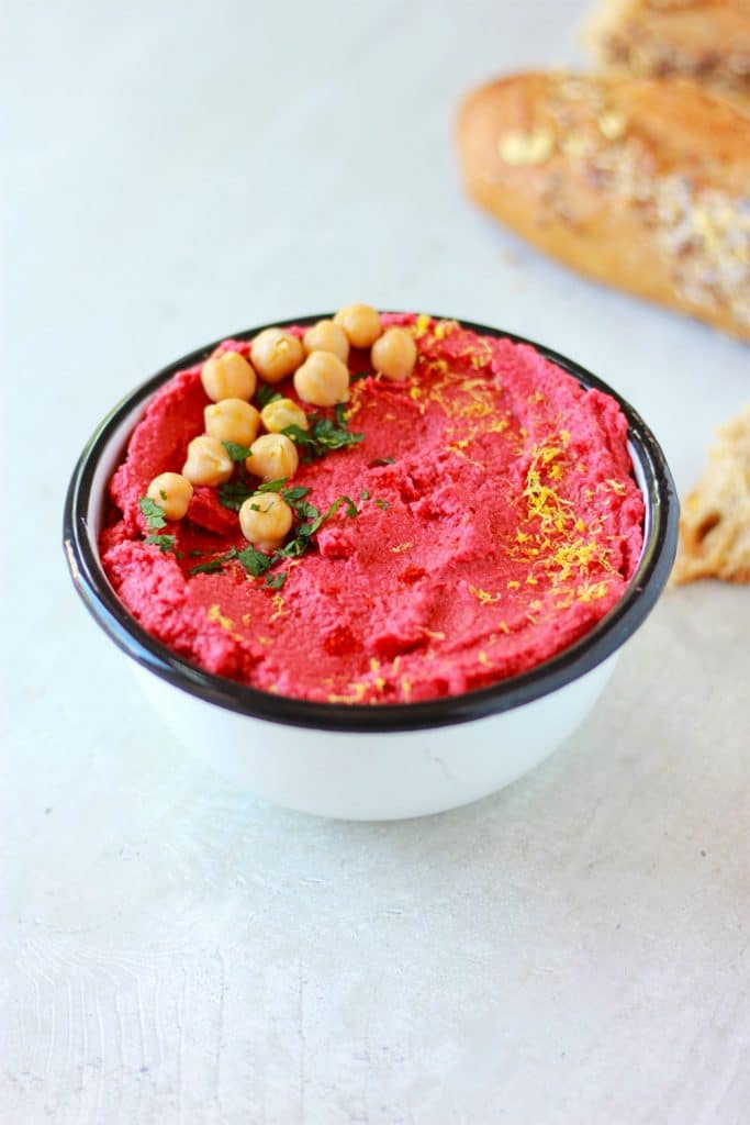 Red spicy beetroot hummus in a white bowl with chickpeas, chopped mint and lemon zest on top.