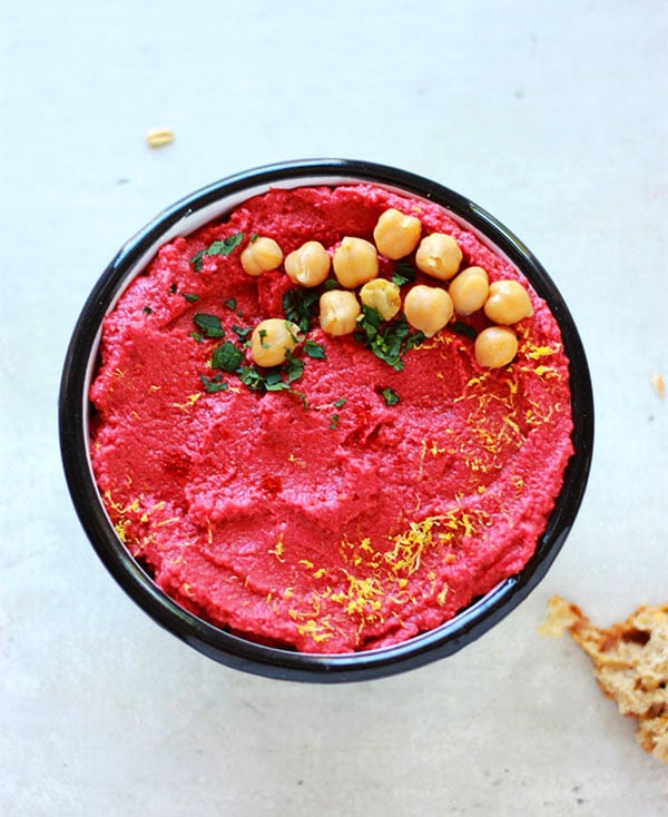 Overhead view of red spicy beetroot hummus in bowl with lemon zest, chickpeas and chopped mint on top.