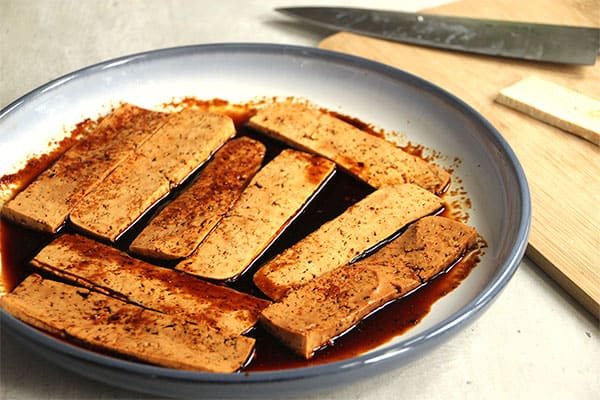 Tofu strips marinated in soy sauce on blue plate with knife in background.
