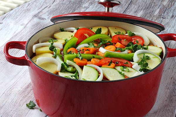 Sliced potatoes, onions, green pepper, carrots, zucchini, diced celery and fresh parsley are arranged in a large red baking pot.