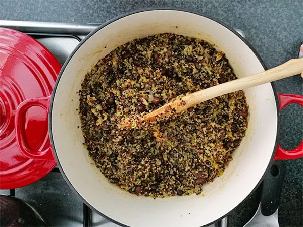 quinoa and leeks and black beans are cooking in a red pot with a wooden spoon on the stovetop.