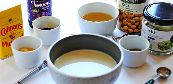 small gray bowl of plant milk, small dish of dry mustard powder with can, dish of Tamari, aqufaba with chickpea can and kelp powder in small dish.