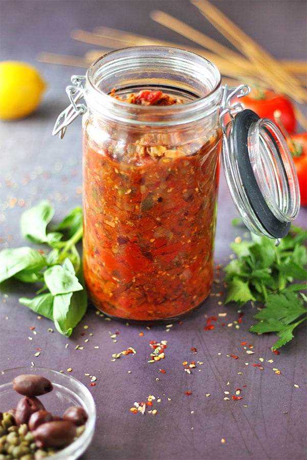 Make healthy Puttanesca sauce with tomatoes, kalamata olives, capers, basil, parsley, oregano, onions, garlic and red chili flakes in tall glass mason jar with olives and capers in foreground.