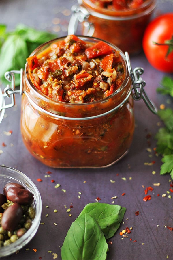 Make healthy Puttanesca sauce with tomatoes, kalamata olives, capers, basil, parsley, oregano, onions, garlic and red chili flakes in short, glass mason jar with another in background.