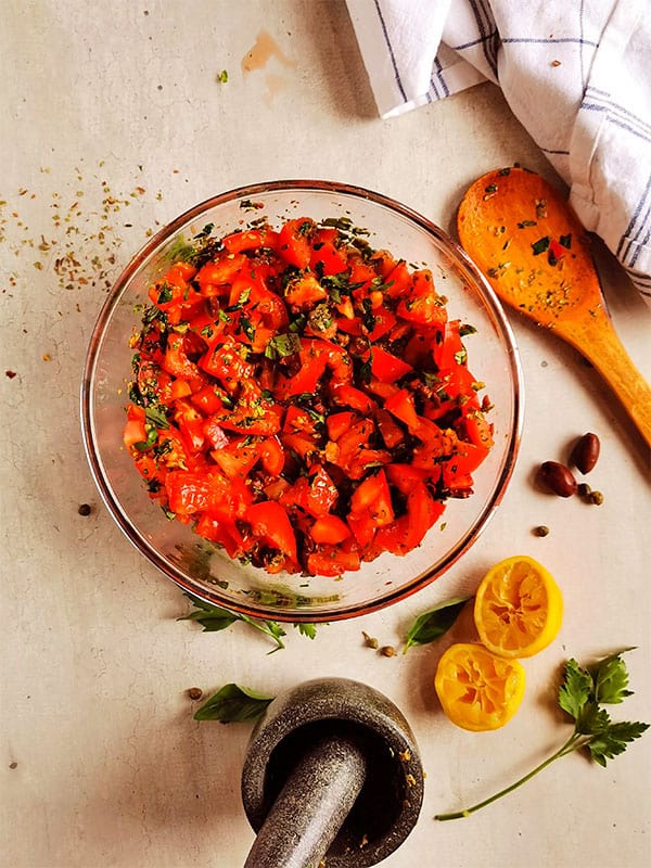 overhead shot of diced tomatoes, fresh parsley and basil, oregano, kalamata olives, capers and lemon juice on board with pestle and mortar and squeezed lemon halves.