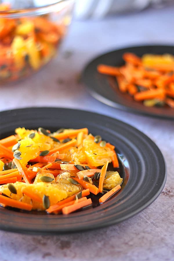 carrot, orange cumin pepita slaw on black plates with glass bowl of slaw in background.