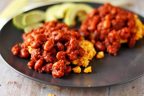 2 corn fritters are smothered with dark red baked chipotle beans on black plate with avocado and lime slices.