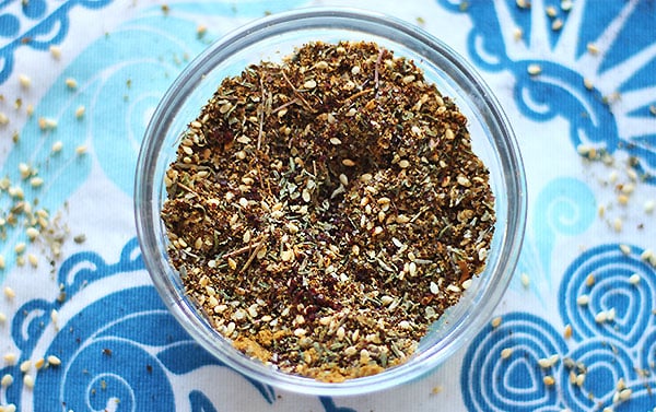 Za'atar spice blend of toasted sesame seeds, thyme, marjoram, cumin, sumac, salt and pepper in small, clear dish on blue cloth.