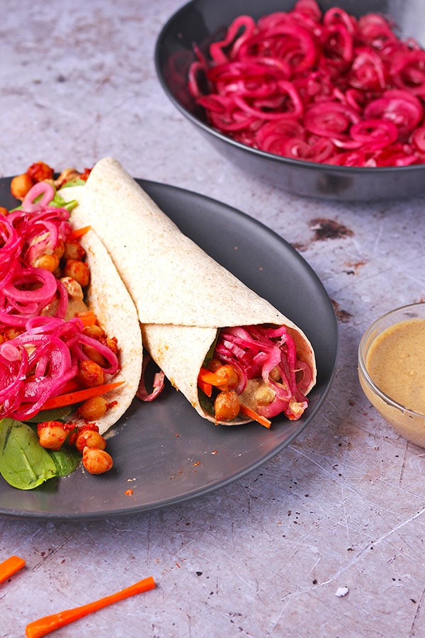 BBQ chickpea wraps with marinated red onions and creamy tahini dressing with sliced carrots and spinach leaves on black plate with open wrap and bowl of onions and dressing. .