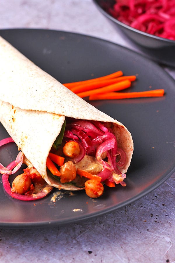 Front view of BBQ chickpea wraps with marinated red onions and creamy tahini dressing with sliced carrots and spinach leaves on black plate.