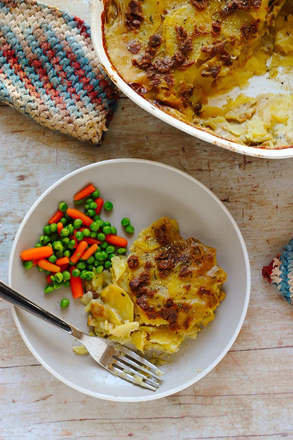 vegan scalloped potatoes with sheet pan bacon on plate with fork and peas and carrots with baking dish of more scalloped potatoes.