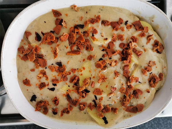 vegan scalloped potatoes are topped with sheet pan bacon bits and placed in a white baking dish ready for the oven.