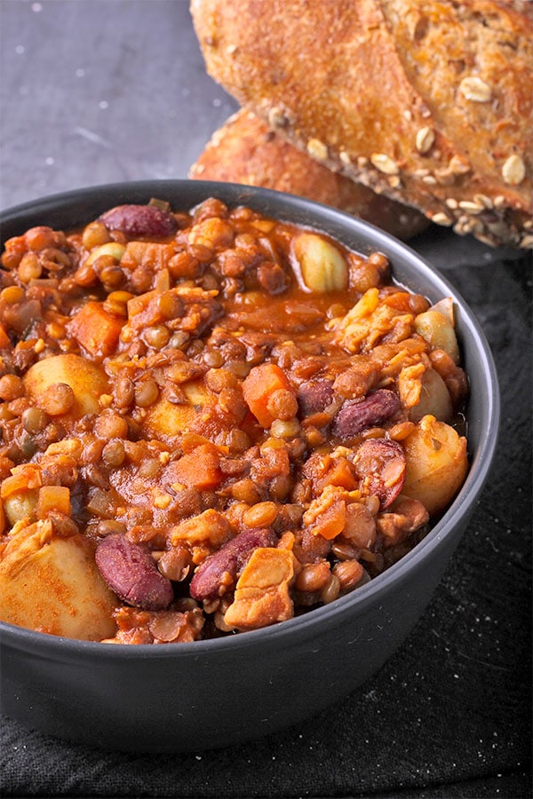 Closeup of loaded tempeh stew with tempeh, kidney beans, fava beans, lentils, potatoes, carrots and tomato sauce in black bowl.