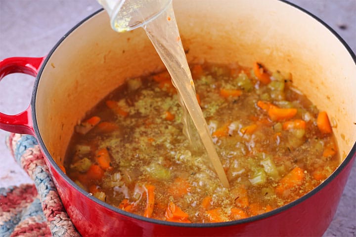 Pouring vegetable broth into red soup pot with carrots, onions, celery and garlic