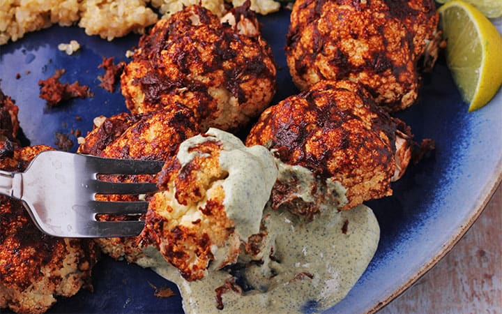 Roasted BBQ cauliflower on blue plate is dipped into pepita dressing with fork.