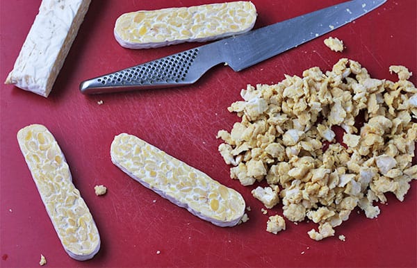 tempeh block is cut into strips and crumbled on red mat.