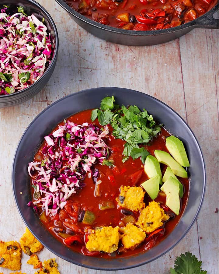 overhead shot of black bean chili with jalapeno polenta squares, cabbage slaw, chopped cilantro and avocado slices in black bowl