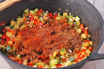 Mixed spices are added to diced onions, zucchini, red pepper and garlic in black pot