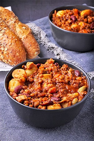 Loaded tempeh stew with kidney beans, fava beans, potatoes, lentils and tomato sauce in black bowls with bread.