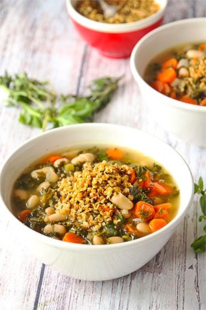 Italian white bean soup with carrots, kale and fresh herbs in white bowl with hemp Parmesan on top.