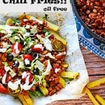 loaded chili fries with red chili, lettuce, tomatoes, pickled jalapenos, black olives, cilantro and vegan sour cream on plate with parchment paper, fries on brown board and black pot of chili with recipe title in black text..