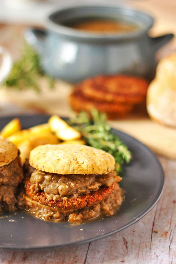 Sweet potato biscuits are loaded with Tempeh sausage patties and smothered in lentil gravy on a black plate with blue pot of gravy, biscuits and sausage patties in background.