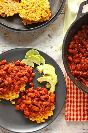 overhead shot of 2 corn fritters smothered with red, chipotle baked beans on black plate with avocado and lime slices and black pan of corn fritters and black pan of chipotle beans on red cloth.