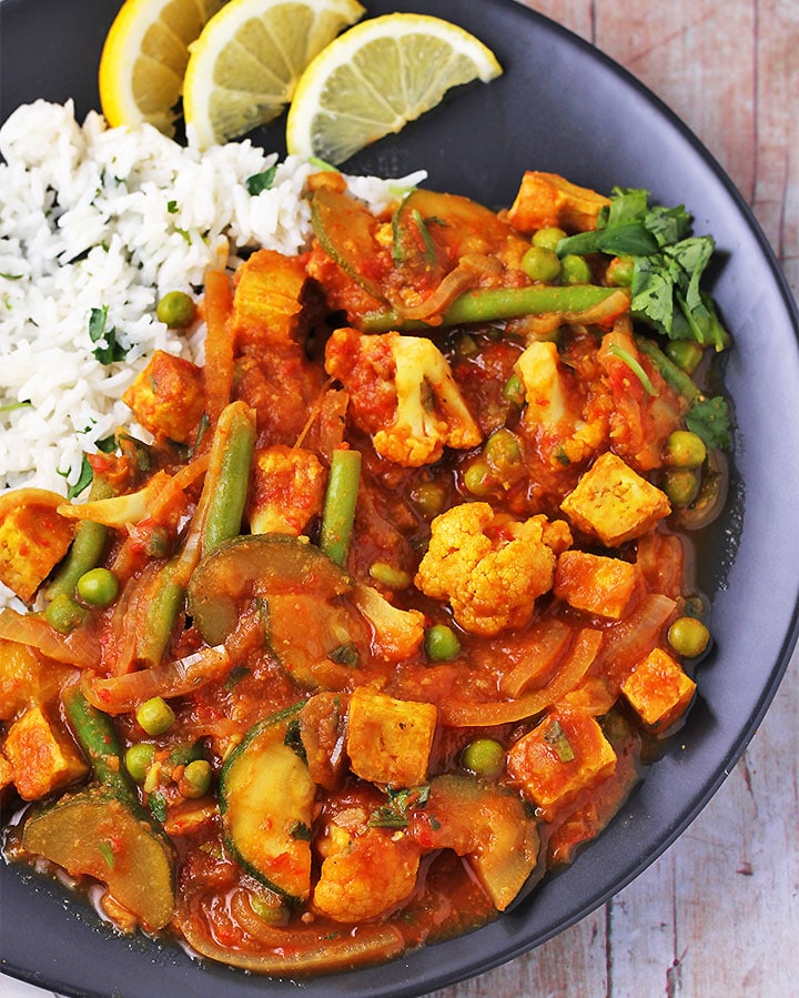 Tomato vegetable curry with zucchini, green beans, peas, onions and cauliflower and crispy tofu on black plate with rice