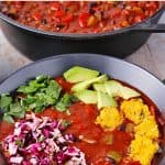 Bowl of black bean chili with cocoa powder topped with jalapeno polenta squares, cabbage slaw, chopped cilantro and avocado cubes with black pan of chili in background