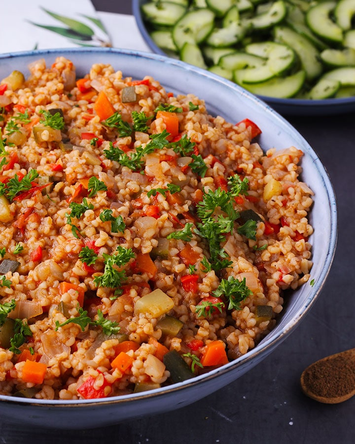Close up with bulgur and vegetables in blue bowl with wooden spoon of Baharat spices and cucumber-mint salad