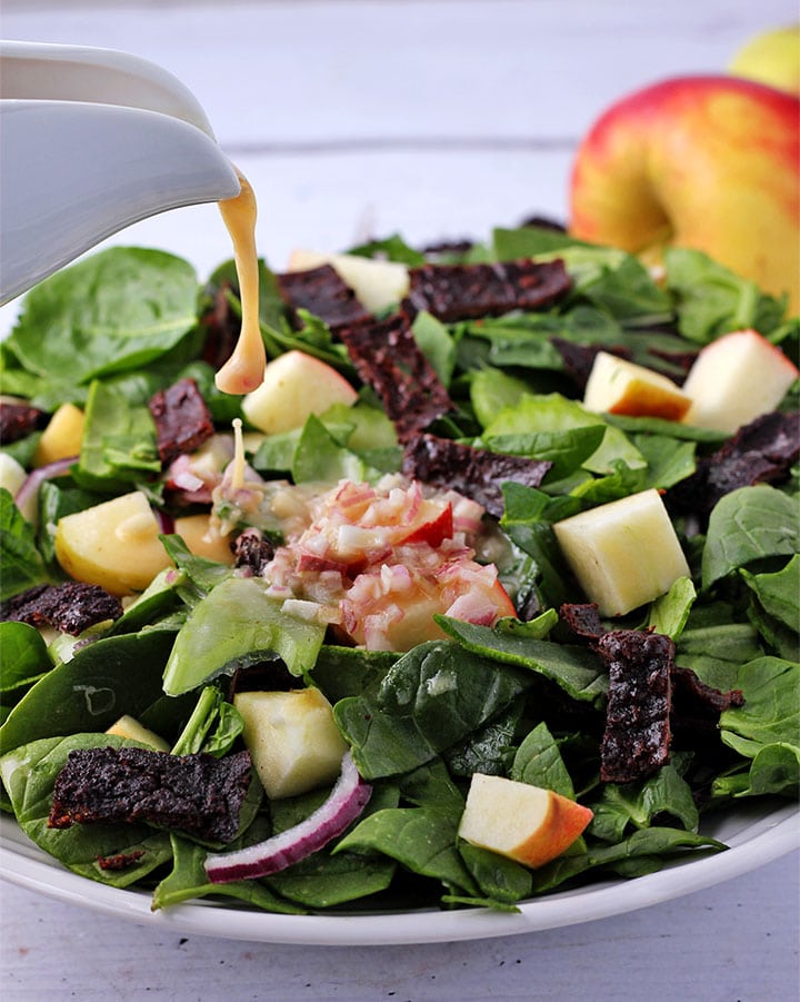 Dressing with mustard and, red wine vinegar and shallots is poured on salad with spinach, red onions, apple cubes and plant-based bacon