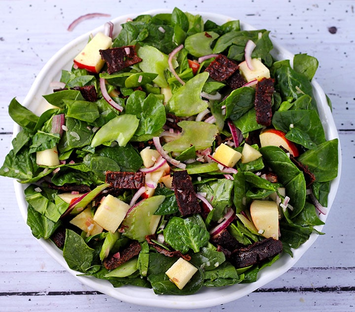 overhead shot of spinach apple salad with plant-based bacon and red onion in white bowl.