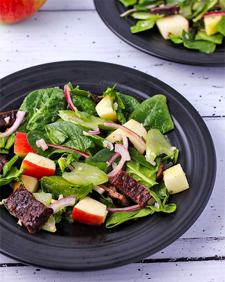 Spinach apple salad with plant-based bacon, red onion and celery on black plate