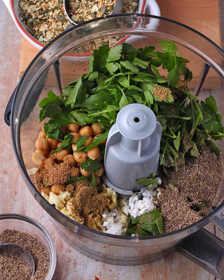 Falafel ingredients including chickpeas, ground chia seeds, garlic, parsley, cilantro, baking soda, cumin and coriander are placed in a food processor.