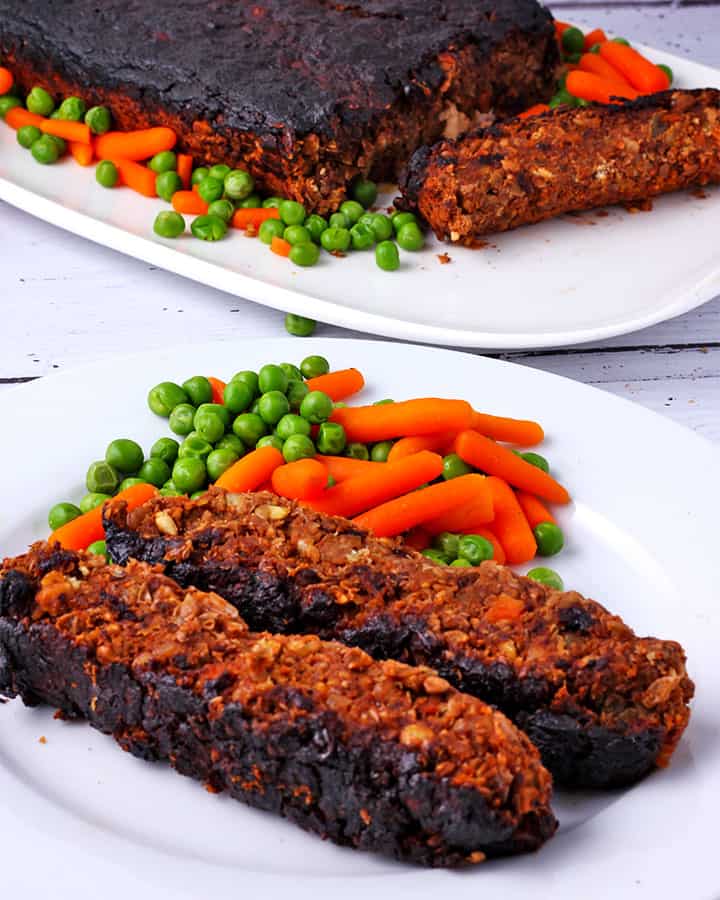 2 slices of baked lentil loaf on white plate with cooked baby carrots and peas and big serving plate with lentil loaf and veggies.