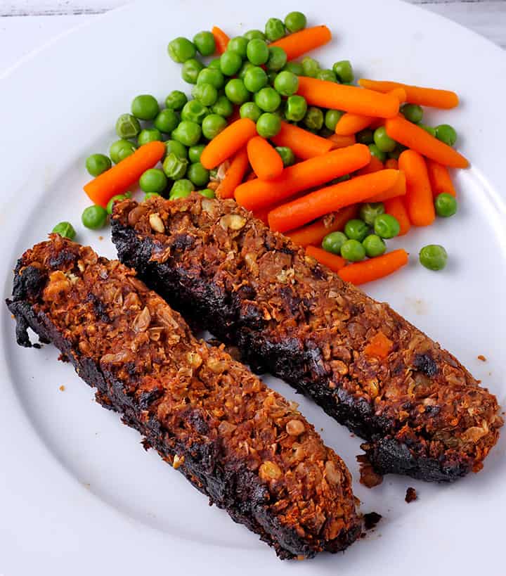 overhead shot of 2 slices of baked lentil loaf with cooked baby carrots and peas on white plate and white serving plate with more loaf slices and loaf and cooked veggies on it.
