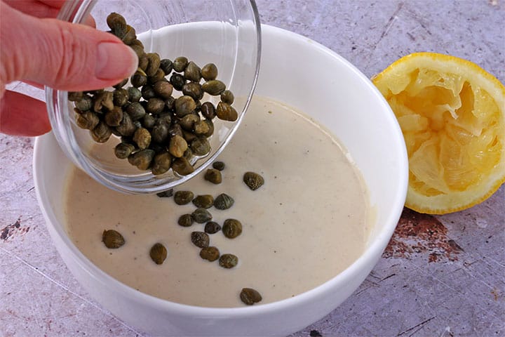 green capers are added to white bowl of almond Caesar salad dressing