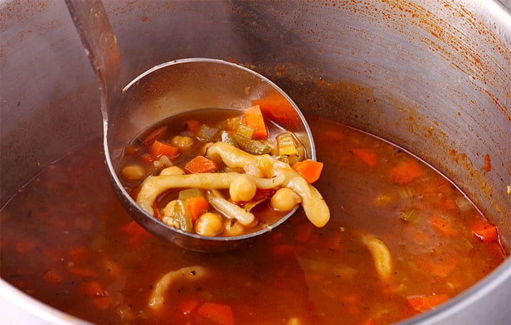 ladle filled with chickpea soup, homemade noodles, carrots, onions and celery is held over soup pot filled with soup.