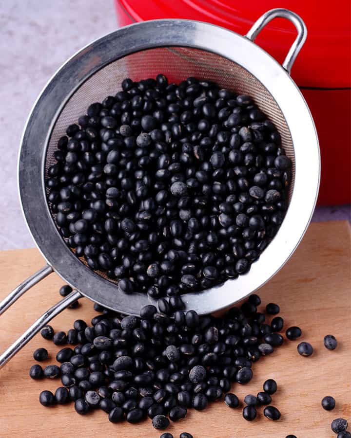 Dried black beans are rinsed and placed in a strainer that is tilted so some of the beans spill onto a wooden board.