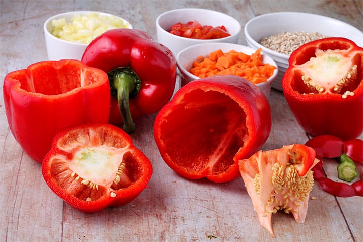 4 whole red bell peppers with tops cut off and cores on board with bowls of chopped onions, carrots and uncooked barley.