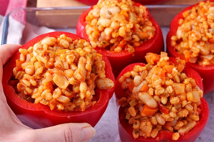 Whole red pepper stuffed with barley and white beans and tomato sauce is placed in glass baking dish with hand.