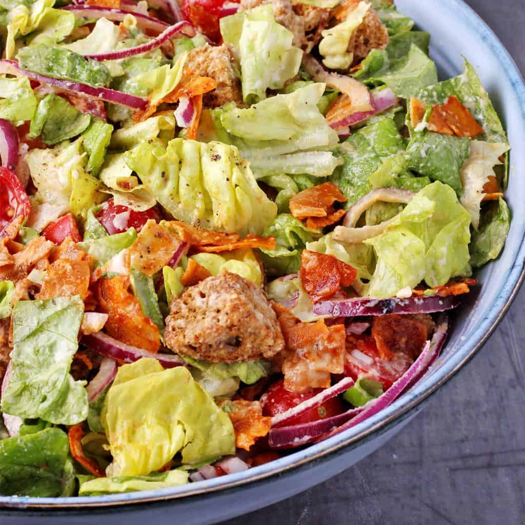 close up picture of vegan Caesar salad with chopped lettuce, sliced cherry tomatoes, sliced red onion, coconut bacon, baked croutons and almond dressing